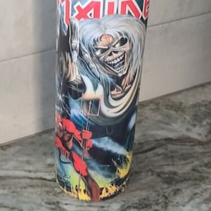 May include: A tall, stainless steel tumbler with a black lid. The tumbler features a colorful graphic of a metal band, with the band's name "IRON MAIDEN" printed at the top. The graphic includes a red, white, and black image of a man with long white hair and a red and black face.
