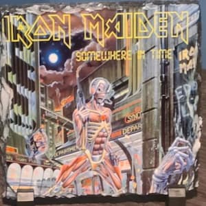May include: A black slate coaster with a colorful image of the band Iron Maiden. The image features the band's name in yellow letters and the album title "Somewhere in Time" in white letters. The image also features a skeletal figure with a red star on its chest.