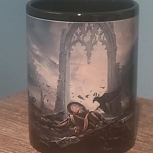 May include: Black ceramic mug featuring a detailed fantasy scene. The artwork depicts a fallen figure in tattered clothing, a gothic archway, and ominous crows against a stormy sky. The mug has a black interior and rim.