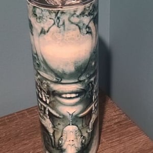 May include: A stainless steel tumbler with a white and green abstract design featuring a face and hands. The design is reminiscent of a creature from a science fiction movie.