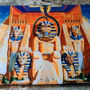 May include: A colorful painting of a pharaoh sitting on a throne with a snake above him. The pharaoh is surrounded by four smaller figures with animal heads. The painting is set against a background of a blue sky with clouds.