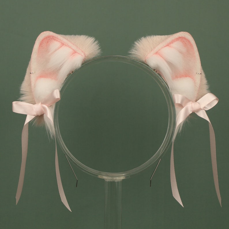 May include: A pair of pink furry cat ears with white tips and pink satin bows attached to a clear plastic headband.