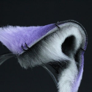May include: Purple and white faux fur cat ears headband with black metal clips. The headband is made of soft, plush fabric and is perfect for adding a touch of whimsy to any outfit.