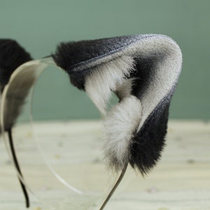 May include: A black and white furry headband with cat ears. The headband is made of soft, plush fabric and is perfect for adding a touch of whimsy to any outfit.