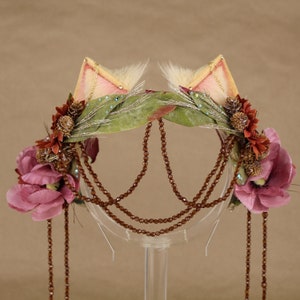 May include: A pink and brown floral headband with cat ears, featuring a leafy green base, brown beads, and pink flowers.