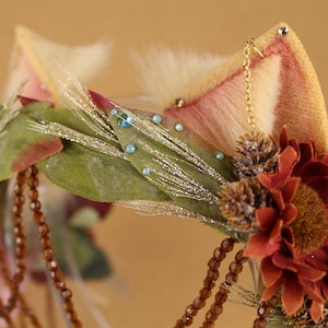 May include: A brown, green, and white floral headband with a pink and white fur ear on the right side. The headband is decorated with small blue rhinestones and brown beads.