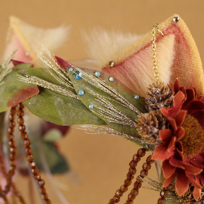 May include: A brown, green, and white floral headband with a pink and white fur ear on the right side. The headband is decorated with small blue rhinestones and brown beads.