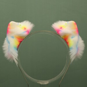 May include: A white headband with two fluffy rainbow-colored cat ears attached. The headband is made of a thin gold wire.