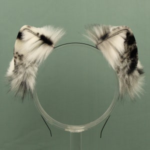 May include: A pair of white and black faux fur cat ears with a black headband. The ears are fluffy and have a spotted pattern.
