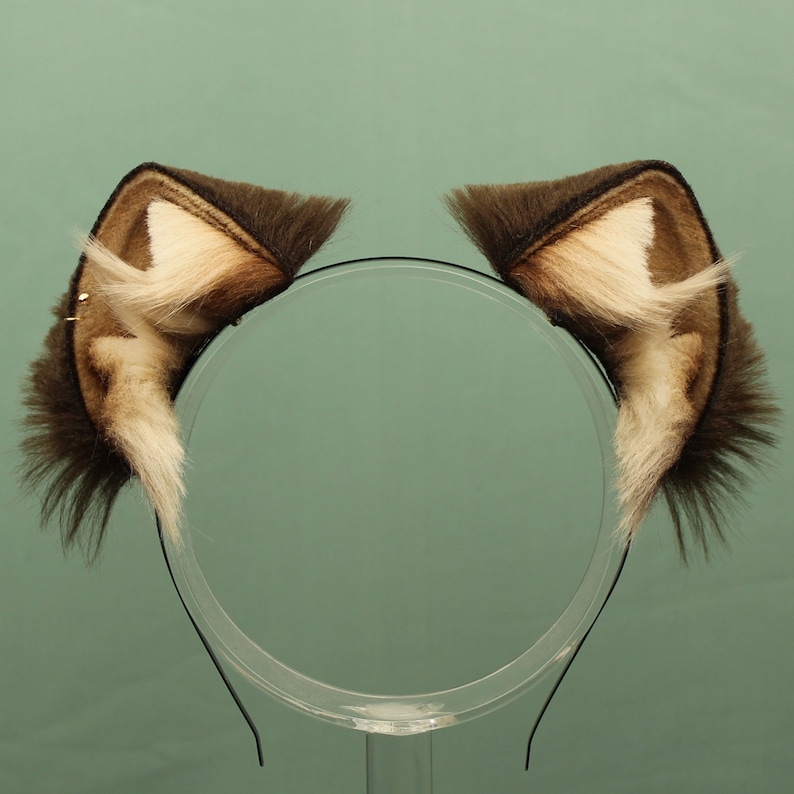 May include: A pair of brown and white faux fur cat ears on a black headband. The headband is on a clear stand.