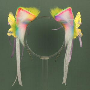May include: A pair of rainbow cat ears with yellow butterfly wings and purple and silver ribbons. The ears are made of faux fur and have a white base.
