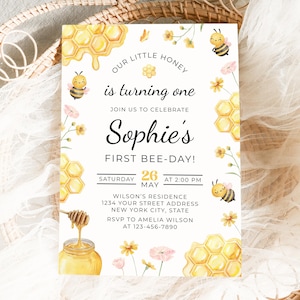 Può includere: Un invito ad acquerello a tema api. Il testo recita "Our little honey is turning one" e "Sophie's First Bee-Day!" Favo, api e fiori decorano l'invito. La data è il 26 maggio alle 14:00.