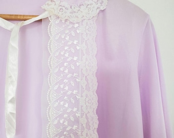 Puede incluir: Una prenda de manga larga en color lavanda claro que presenta un cuello de encaje blanco y un panel vertical de encaje blanco en la parte delantera. La pieza incluye un lazo de cinta blanca en el cuello y cuelga de una percha de madera.