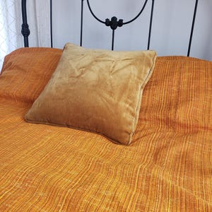 May include: A bed with an orange textured bedspread and a mustard-colored pillow. The bed has a black wrought iron headboard with floral accents. The pillow is square and appears to be made of a soft material, contrasting with the textured bedspread.
