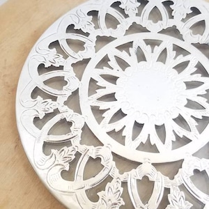 May include: A silver circular trivet with an ornate openwork design. The trivet has a central circular area surrounded by a detailed floral and geometric pattern. The trivet is placed on a light-coloured wooden surface.
