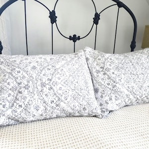 Puede incluir: Dos almohadas decorativas con un estampado floral gris y blanco descansan sobre una colcha crema con tejido de gofre. Las almohadas están frente a un cabecero de hierro forjado negro con detalles florales.