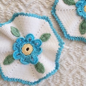 Könnte beinhalten: Zwei weiße Häkeluntersetzer mit einer blauen Blume und grünen Blättern. Die Untersetzer haben einen blau-gewellten Rand. Die Blume hat eine gelbe Mitte. Diese handgefertigten Untersetzer verleihen jedem Zuhause einen floralen Charme.