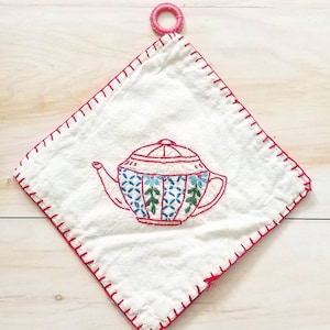 May include: A white cotton tea cosy with red stitching and a hand-embroidered teapot design. The teapot is red, blue, and green with a white background.
