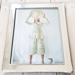 Puede incluir: Un marco de fotos plateado con un paspartú blanco y una foto de una persona que lleva un sombrero blanco y un atuendo estampado en verde y blanco. La foto está sobre un fondo blanco. El texto en la parte inferior de la foto dice "When you think of it that way, it makes the sudden 100th Anniversary seem less strange."