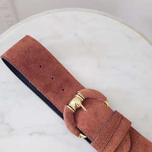 May include: A wide, rust-colored suede belt with a gold-toned buckle. The belt has a black backing and several holes for adjustment. The buckle is a rounded shape with gold accents. The belt is a stylish accessory.
