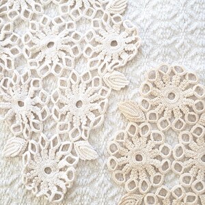May include: Close-up of two cream-colored, handmade crochet doilies with floral and leaf patterns. The doilies are placed on a textured white surface. The intricate design and neutral color palette suggest a vintage or handmade aesthetic.