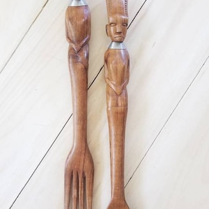Puede incluir: Un juego de servir ensalada de madera con un tenedor y una cuchara. Los mangos están tallados en forma de figuras humanas con anillos de plata alrededor del cuello.