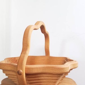 K&ouml;nnte beinhalten: Ein Holzkorb mit einem einzigartigen, gebogenen Griff. Der Korb ist aus hellem braunem Holz gefertigt und hat einen gewellten Rand. Er steht auf einem Holztisch.