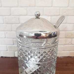 Può includere: Una zuccheriera in vetro trasparente con coperchio e cucchiaio argentati. La ciotola ha un motivo a diamante e poggia su una superficie di legno. Il coperchio ha una piccola sfera sopra.