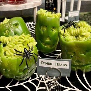 May include: Three green bell peppers carved with facial features and filled with light green rotini pasta. Each pepper is topped with a black plastic spider. A small rectangular sign in front reads "ZOMBIE HEADS" held by a silver metal spiral stand.