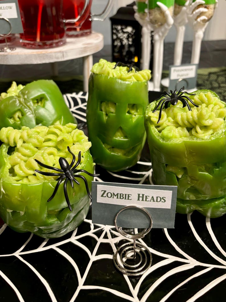 May include: Three green bell peppers carved with facial features and filled with light green rotini pasta. Each pepper is topped with a black plastic spider. A small rectangular sign in front reads "ZOMBIE HEADS" held by a silver metal spiral stand.