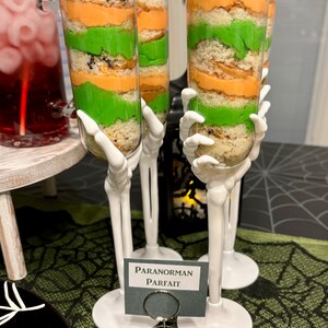 May include: Two clear plastic dessert cups filled with layers of orange and green cake and frosting, topped with green sprinkles. The cups are held by white plastic skeleton hand stems and bases. A small sign reads PARANORMAN PARFAIT.