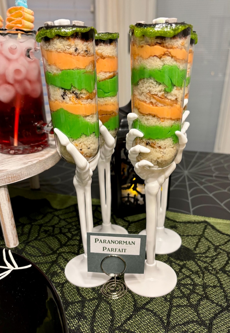 May include: Two clear plastic dessert cups filled with layers of orange and green cake and frosting, topped with green sprinkles. The cups are held by white plastic skeleton hand stems and bases. A small sign reads PARANORMAN PARFAIT.