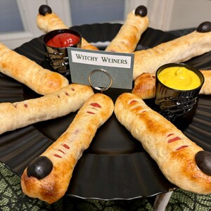 May include: A platter of finger-shaped hot dogs wrapped in golden-brown dough, each topped with a black olive fingernail. The dish includes small cups of red ketchup and yellow mustard, with a sign reading Witchy Weiners in the center.