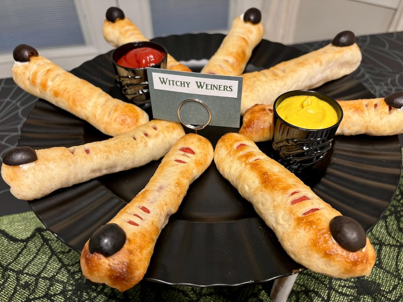 May include: A platter of finger-shaped hot dogs wrapped in golden-brown dough, each topped with a black olive fingernail. The dish includes small cups of red ketchup and yellow mustard, with a sign reading Witchy Weiners in the center.