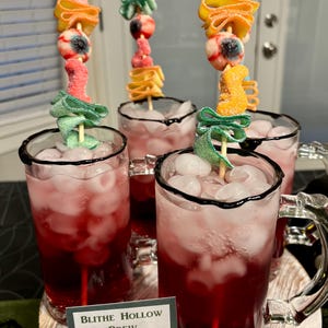 May include: Four glass mugs filled with red liquid and ice cubes, each featuring a skewer topped with gummy candies including an eyeball, green ribbons, and orange shapes. A small sign in front reads Blithe Hollow Brew in black text.