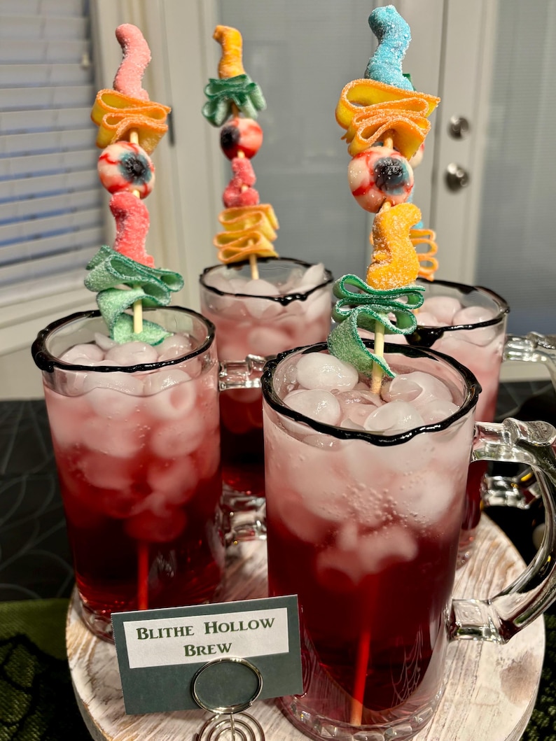 May include: Four glass mugs filled with red liquid and ice cubes, each featuring a skewer topped with gummy candies including an eyeball, green ribbons, and orange shapes. A small sign in front reads Blithe Hollow Brew in black text.
