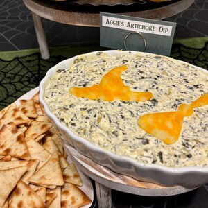 May include: A white ceramic scalloped baking dish filled with creamy spinach and artichoke dip, topped with two orange cheese cutouts shaped like a witch hat and a broom. A small sign reads Aggie's Artichoke Dip, accompanied by triangle crackers.