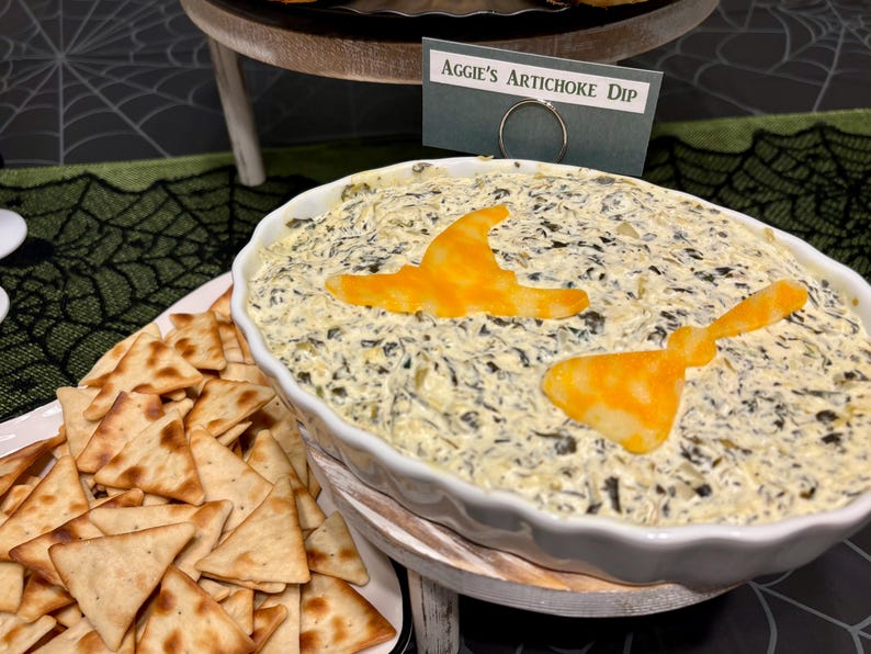 May include: A white ceramic scalloped baking dish filled with creamy spinach and artichoke dip, topped with two orange cheese cutouts shaped like a witch hat and a broom. A small sign reads Aggie's Artichoke Dip, accompanied by triangle crackers.