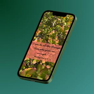 Könnte beinhalten: Ein grünes Smartphone zeigt ein Bild mit Naturmotiv und dem Text "Ich vermag alles durch den, der mich stark macht." aus Philipper 4,13. Der Hintergrund zeigt grüne und rot gefärbte Pflanzenblätter.
