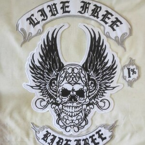 Puede incluir: Un parche blanco y negro con un cráneo alado, el texto "LIVE FREE" arriba y "RIDE FREE" abajo. También se incluye un pequeño parche con "1%". El diseño es de estilo gótico.