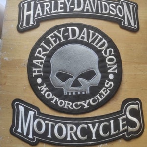 May include: Three Harley-Davidson motorcycle patches. The top patch is a black banner with white stitching that reads "Harley-Davidson". The middle patch is a black circle with white stitching that reads "Harley-Davidson Motorcycles" and features a silver skull with black eye sockets. The bottom patch is a black banner with white stitching that reads "Motorcycles".