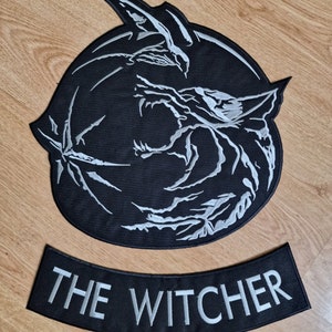 May include: A black and silver embroidered patch with a wolf's head design and the text "THE WITCHER" in a silver font.