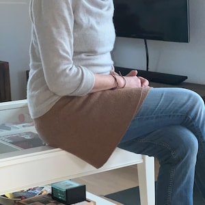 Könnte beinhalten: Ein hellgrauer Pullover mit einem Wasserfallausschnitt und langen Ärmeln. Ein brauner Rock und blaue Jeans sind ebenfalls zu sehen. Die Person sitzt auf einem weißen Tisch. Ein schwarzer Fernseher ist im Hintergrund.