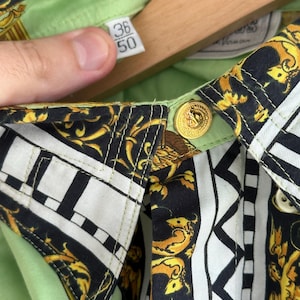 K&ouml;nnte beinhalten: Nahaufnahme eines Hemdes mit Knopfleiste und lebhaftem Muster. Das Hemd hat ein schwarzes und goldenes Barockdesign, akzentuiert mit wei&szlig;en geometrischen Formen. Der Kragen und das Innenfutter sind hellgr&uuml;n. Das Hemd hat goldene Kn&ouml;pfe und ein Etikett mit den Zahlen 36 und 50.