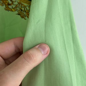 K&ouml;nnte beinhalten: Nahaufnahme eines hellgr&uuml;nen Stoffes, m&ouml;glicherweise Seide oder ein &auml;hnliches Material, gehalten von einer Hand. Der Stoff hat eine glatte Textur und einen leichten Glanz. Im Hintergrund ist ein Abschnitt eines gemusterten Stoffes mit goldenen und orangenen Akzenten zu sehen.