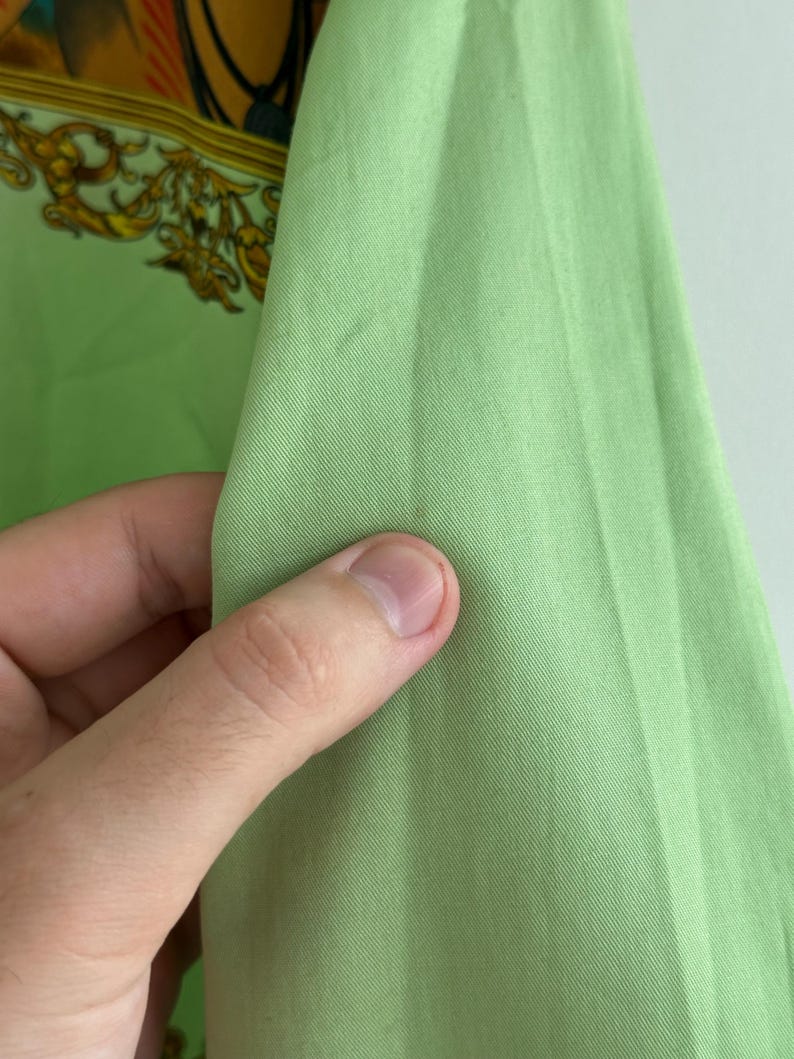 K&ouml;nnte beinhalten: Nahaufnahme eines hellgr&uuml;nen Stoffes, m&ouml;glicherweise Seide oder ein &auml;hnliches Material, gehalten von einer Hand. Der Stoff hat eine glatte Textur und einen leichten Glanz. Im Hintergrund ist ein Abschnitt eines gemusterten Stoffes mit goldenen und orangenen Akzenten zu sehen.