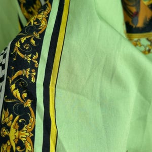 K&ouml;nnte beinhalten: Nahaufnahme eines hellgr&uuml;nen Stoffes mit dekorativem Rand. Der Rand weist schwarze und goldene Barockmotive mit gelben und schwarzen Streifen auf. Im Hintergrund ist ein Portr&auml;t einer Person zu sehen.