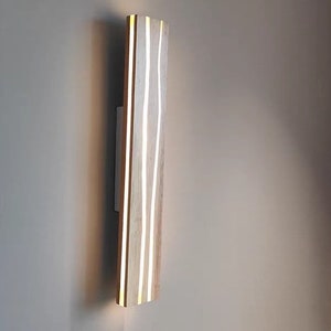 May include: A wooden wall sconce with a light wood finish and white lines running vertically. The sconce is mounted on a wall.