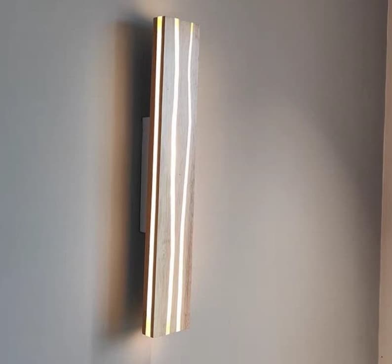 May include: A wooden wall sconce with a light wood finish and white lines running vertically. The sconce is mounted on a wall.