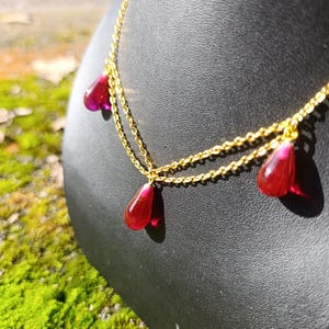 May include: A gold-colored necklace with two delicate chains. The necklace features three teardrop-shaped, deep red pendants. The necklace is displayed on a black mannequin, with a blurred green and brown background.
